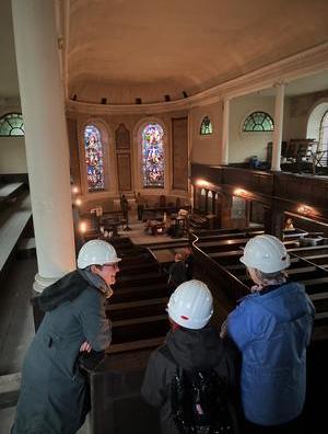 3 people wearing hard hats looking down from the upper gallery at a group of people cleaning the nave of the church 